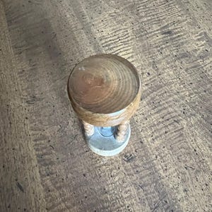 K&ouml;nnte beinhalten: Eine kleine h&ouml;lzerne Sanduhr mit einer hellbraunen, abgerundeten Oberseite und einem hellgrauen Sockel. Die Sanduhr hat eine hellbraune Holzmitte mit zwei beigefarbenen, seilartigen St&uuml;tzen.