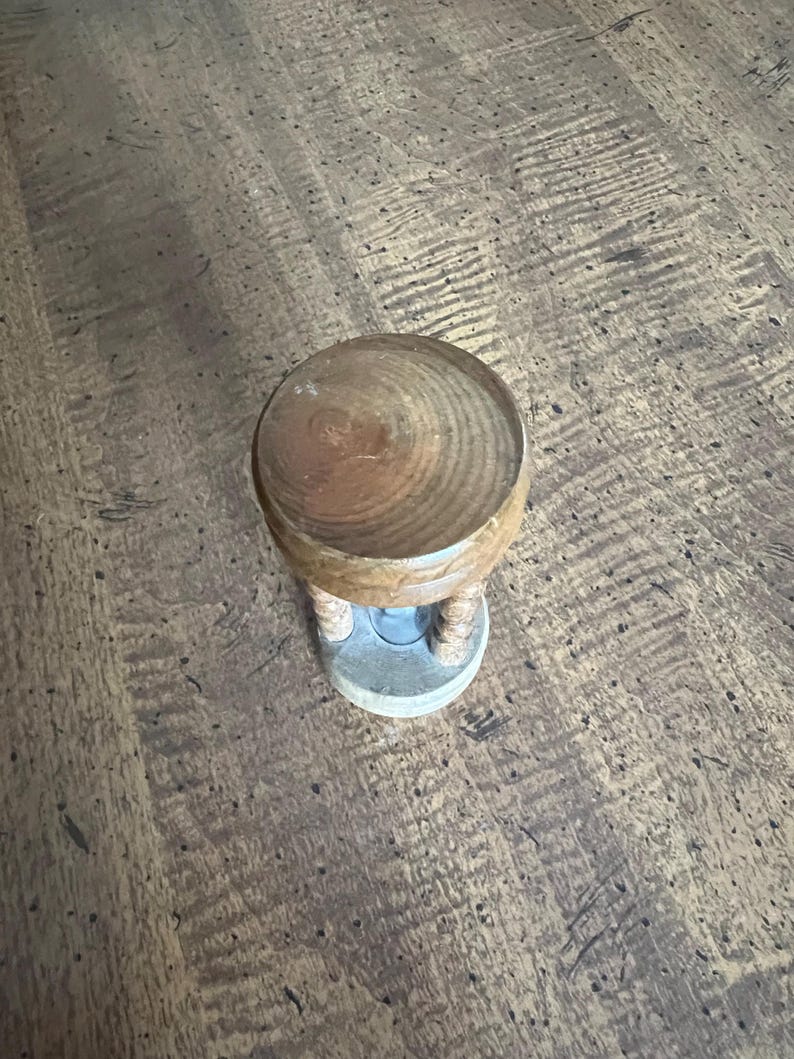 K&ouml;nnte beinhalten: Eine kleine h&ouml;lzerne Sanduhr mit einer hellbraunen, abgerundeten Oberseite und einem hellgrauen Sockel. Die Sanduhr hat eine hellbraune Holzmitte mit zwei beigefarbenen, seilartigen St&uuml;tzen.