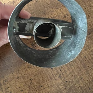 May include: A vintage, circular metal cookie cutter with a central tube and a handle. The cutter is made of a dull, silver-colored metal and shows signs of wear, suggesting it's an antique kitchen tool. The design is simple and functional.