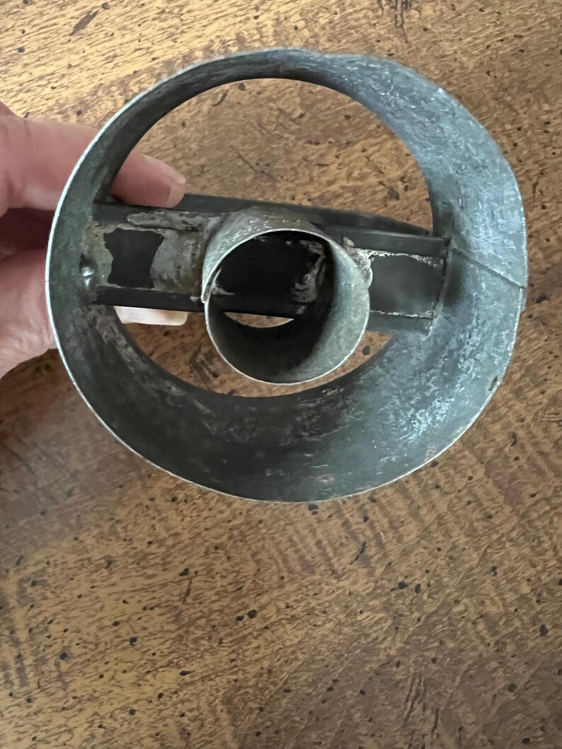 May include: A vintage, circular metal cookie cutter with a central tube and a handle. The cutter is made of a dull, silver-colored metal and shows signs of wear, suggesting it's an antique kitchen tool. The design is simple and functional.