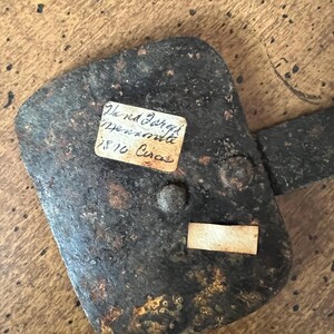 May include: A weathered, dark metal hinge with a rectangular plate and a handle. Two small paper labels are attached, one with handwritten text. The hinge is riveted and shows signs of age and rust, set against a wooden surface.
