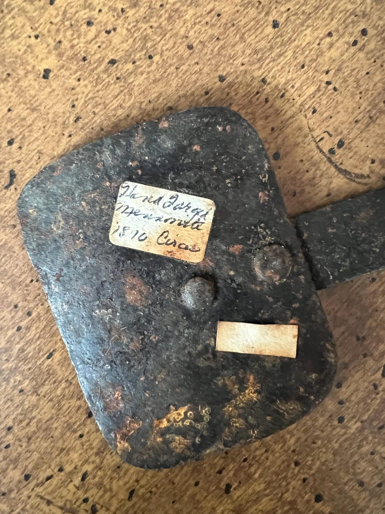 May include: A weathered, dark metal hinge with a rectangular plate and a handle. Two small paper labels are attached, one with handwritten text. The hinge is riveted and shows signs of age and rust, set against a wooden surface.