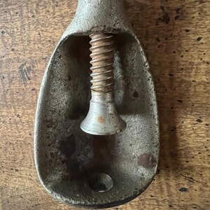 May include: Antique metal nutcracker with a screw mechanism. The nutcracker is made of a weathered, silver-toned metal with a teardrop-shaped bowl and a central screw. The screw is rusty, and the metal shows signs of age and use.