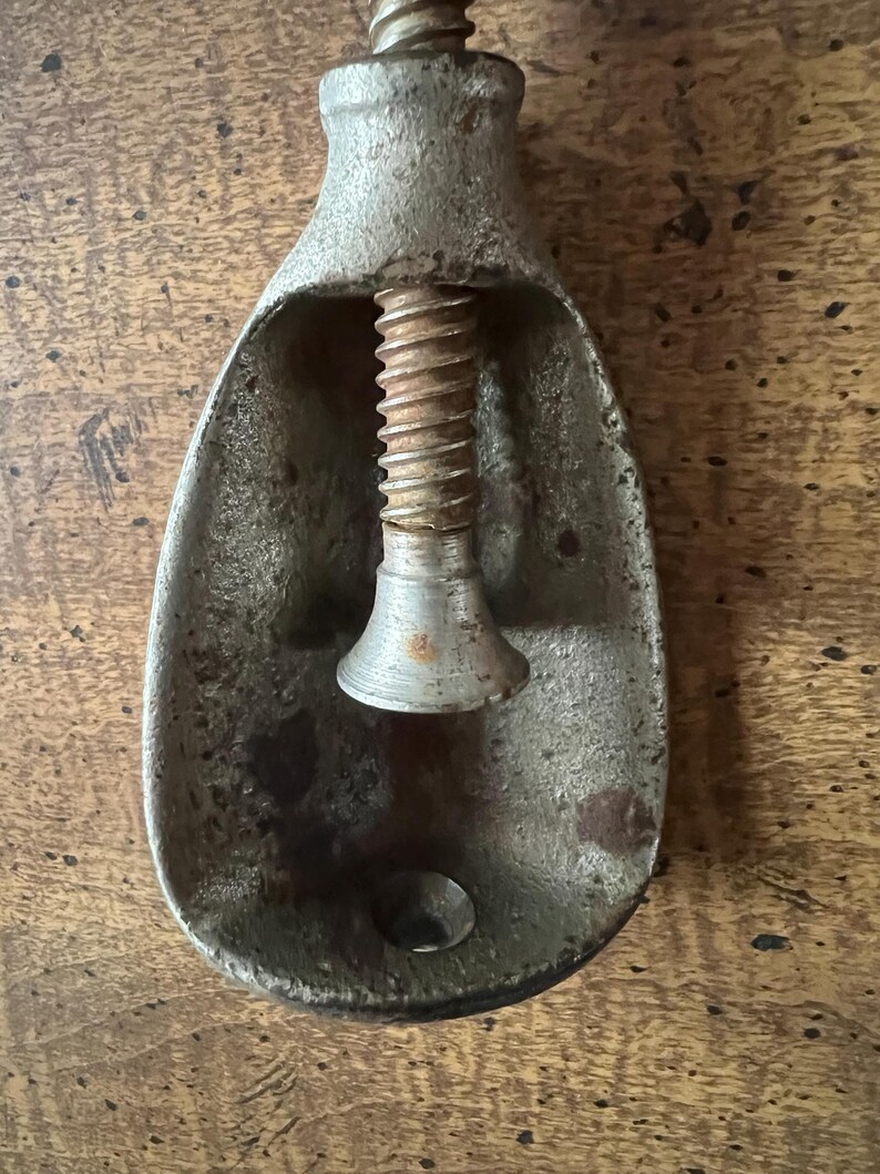 May include: Antique metal nutcracker with a screw mechanism. The nutcracker is made of a weathered, silver-toned metal with a teardrop-shaped bowl and a central screw. The screw is rusty, and the metal shows signs of age and use.