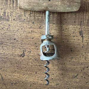 May include: A vintage corkscrew with a wooden handle and a metal screw. The handle is a light brown color, and the metal components show signs of age. The corkscrew is designed for removing corks from wine bottles.