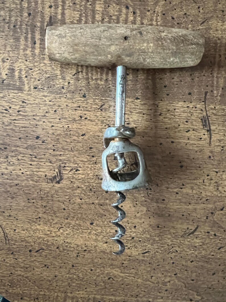 May include: A vintage corkscrew with a wooden handle and a metal screw. The handle is a light brown color, and the metal components show signs of age. The corkscrew is designed for removing corks from wine bottles.