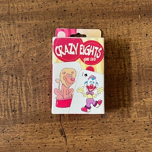 May include: A box of Crazy Eights game cards with a pink, yellow, and white design. The box features a cartoon lion and a clown.