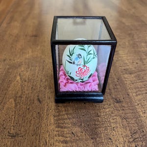 May include: A hand-painted egg with a bird and floral design is displayed in a glass case with a pink satin cushion.