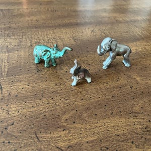 May include: Three small elephant figurines in different colors. One is teal, one is silver, and the smallest is gray and white. The elephants are posed on a brown wooden surface.