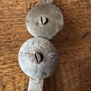 May include: Antique metal nutcracker with two circular, riveted plates connected by a handle. The top plate has a small tab. The bottom plate is stamped with "RODGERS ENGLAND" and a cross symbol. The metal has a worn, aged appearance.