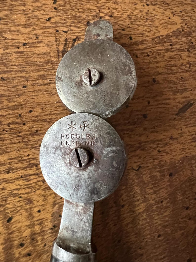 May include: Antique metal nutcracker with two circular, riveted plates connected by a handle. The top plate has a small tab. The bottom plate is stamped with "RODGERS ENGLAND" and a cross symbol. The metal has a worn, aged appearance.