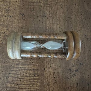 K&ouml;nnte beinhalten: Eine h&ouml;lzerne Sanduhr mit hellem Sand im Inneren. Die Sanduhr hat eine zylindrische Form mit abgerundeten Holzenden und einem schmalen Mittelteil. Das Holz hat eine nat&uuml;rliche, hellbraune Oberfl&auml;che.