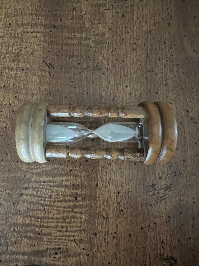 K&ouml;nnte beinhalten: Eine h&ouml;lzerne Sanduhr mit hellem Sand im Inneren. Die Sanduhr hat eine zylindrische Form mit abgerundeten Holzenden und einem schmalen Mittelteil. Das Holz hat eine nat&uuml;rliche, hellbraune Oberfl&auml;che.