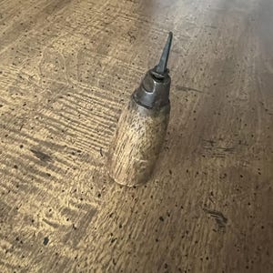 May include: A vintage oil can with a wooden handle and a metal spout. The can has a cylindrical shape and is made of wood and metal. The spout is pointed and angled upwards. The can is resting on a wooden surface.