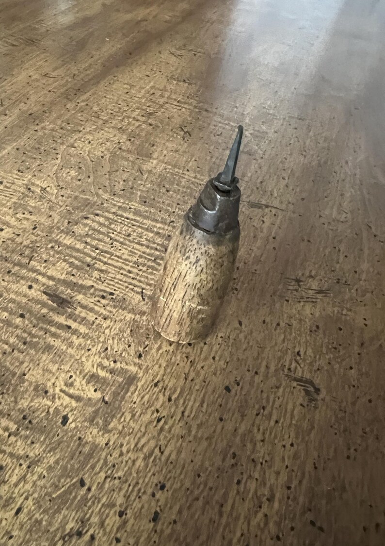 May include: A vintage oil can with a wooden handle and a metal spout. The can has a cylindrical shape and is made of wood and metal. The spout is pointed and angled upwards. The can is resting on a wooden surface.
