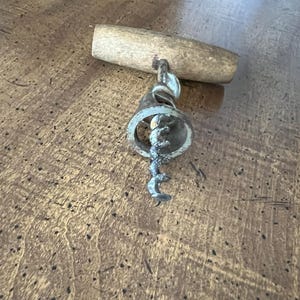 May include: A vintage corkscrew with a wooden handle and a metal screw. The metal components show signs of age and use. The corkscrew is designed for removing corks from wine bottles. The wooden handle is light brown.