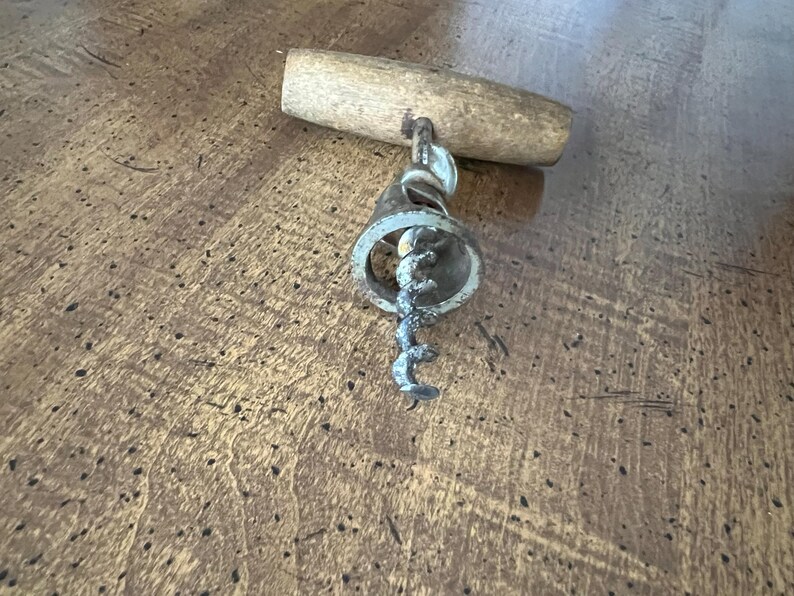 May include: A vintage corkscrew with a wooden handle and a metal screw. The metal components show signs of age and use. The corkscrew is designed for removing corks from wine bottles. The wooden handle is light brown.