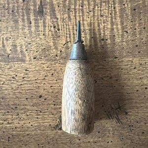 May include: A wooden awl with a metal tip. The awl has a cylindrical wooden handle and a pointed metal tip. The wood has a natural grain pattern. The awl is designed for piercing leather or other materials.