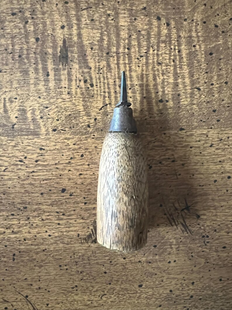 May include: A wooden awl with a metal tip. The awl has a cylindrical wooden handle and a pointed metal tip. The wood has a natural grain pattern. The awl is designed for piercing leather or other materials.