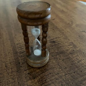 K&ouml;nnte beinhalten: Eine kleine h&ouml;lzerne Sanduhr mit dunkelbraunem Finish. Die Sanduhr hat eine klare Glasmitte, die mit wei&szlig;em Sand gef&uuml;llt ist. Der Holzrahmen hat eine runde Ober- und Unterseite, die durch vier dekorative, gedrehte Beine verbunden sind.