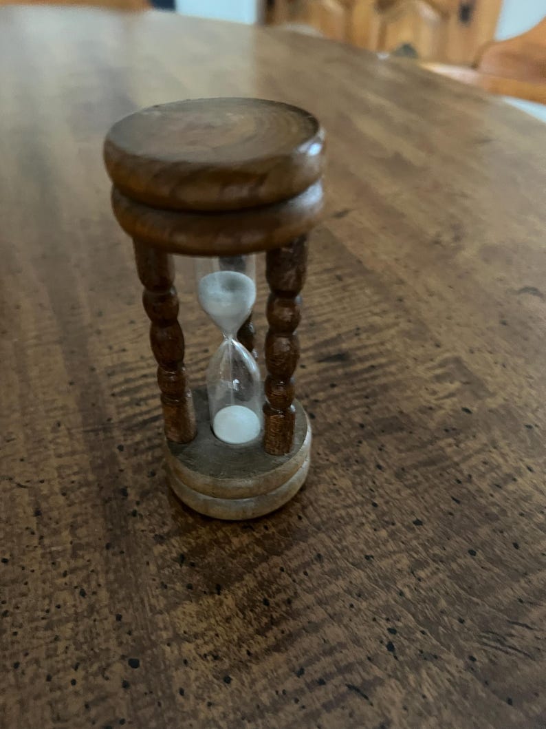 K&ouml;nnte beinhalten: Eine kleine h&ouml;lzerne Sanduhr mit dunkelbraunem Finish. Die Sanduhr hat eine klare Glasmitte, die mit wei&szlig;em Sand gef&uuml;llt ist. Der Holzrahmen hat eine runde Ober- und Unterseite, die durch vier dekorative, gedrehte Beine verbunden sind.
