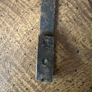 May include: Close-up of a weathered, rectangular metal piece with a riveted end. The metal has a dark, aged appearance with signs of wear and tear, suggesting it is an antique or vintage item. The surface shows a textured pattern.