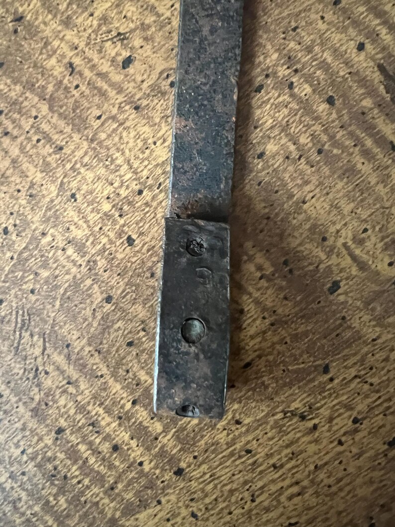 May include: Close-up of a weathered, rectangular metal piece with a riveted end. The metal has a dark, aged appearance with signs of wear and tear, suggesting it is an antique or vintage item. The surface shows a textured pattern.