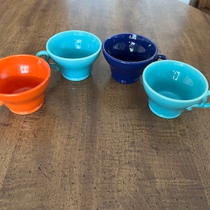 May include: Four ceramic teacups in different colors: orange, blue, light blue, and a darker shade of blue. The cups are arranged in a circle on a wooden table.