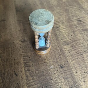 K&ouml;nnte beinhalten: Eine kleine h&ouml;lzerne Sanduhr mit hellblauem Sand. Die Sanduhr hat eine hellbraune Ober- und Unterseite mit einem geschnitzten Holzrahmen. Die Sanduhr ist ein dekorativer Gegenstand.