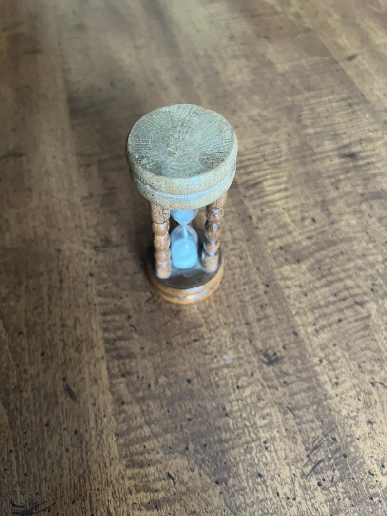 K&ouml;nnte beinhalten: Eine kleine h&ouml;lzerne Sanduhr mit hellblauem Sand. Die Sanduhr hat eine hellbraune Ober- und Unterseite mit einem geschnitzten Holzrahmen. Die Sanduhr ist ein dekorativer Gegenstand.