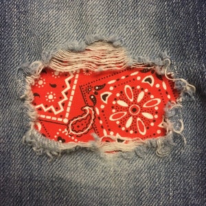 Pode incluir: Um close-up de um par de calças de ganga azuis com um buraco que foi remendado com uma bandana vermelha. A bandana tem um padrão paisley branco.