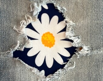 Könnte beinhalten: Nahaufnahme eines weißen Gänseblümchen-Musters auf blauem Hintergrund, das durch ein Loch in einer grauen Jeanshose schaut.
