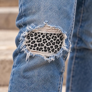 May include: Blue denim jeans featuring a distressed knee area with frayed edges. A fabric patch with a black and white leopard print pattern is visible underneath the ripped section of the denim, showing through the horizontal white threads.