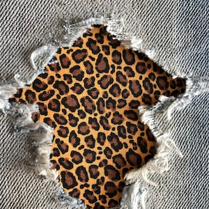May include: A close-up of a ripped pair of blue jeans with a leopard print fabric patch underneath the rip.