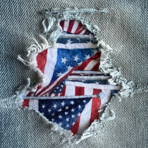 May include: A close-up of a ripped denim fabric with a small hole revealing a printed American flag design. The flag is red, white, and blue with stars and stripes.
