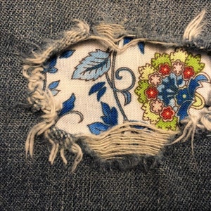 May include: A close-up of a denim fabric with a hole revealing a floral pattern in blue, green, and red colors.