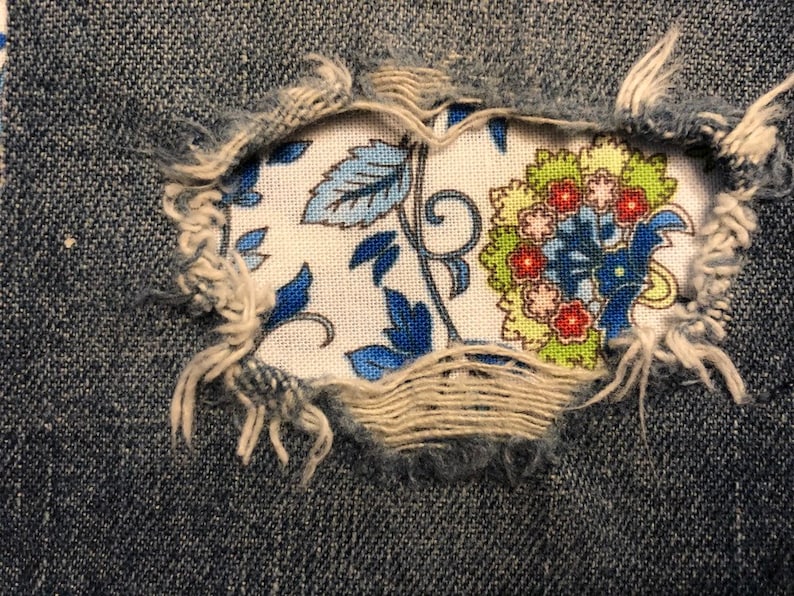 May include: A close-up of a denim fabric with a hole revealing a floral pattern in blue, green, and red colors.