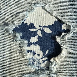 Op de afbeelding: Een close-up van een gescheurde denim stof met een bloemenpatroon dat door het gat zichtbaar is. De denim is lichtblauw en het bloemenpatroon is wit op een donkerblauwe achtergrond.