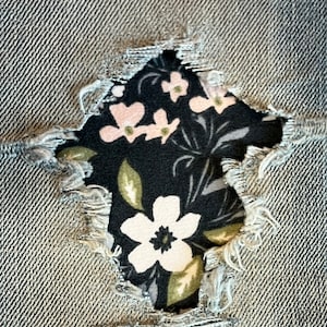May include: A close-up of a pair of denim jeans with a hole revealing a floral fabric underneath. The fabric is black with white and pink flowers.