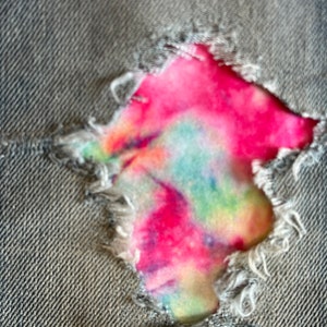 May include: A close-up of a grey denim fabric with a large hole revealing a colourful tie-dye pattern underneath.