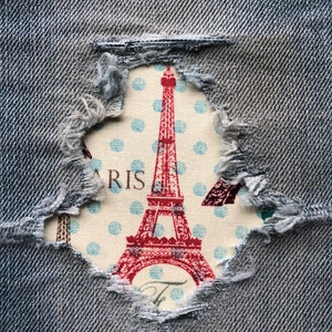 May include: A close-up of a ripped pair of blue jeans with a fabric patch underneath. The patch features a red Eiffel Tower with white polka dots and the word "PARIS" in white text.