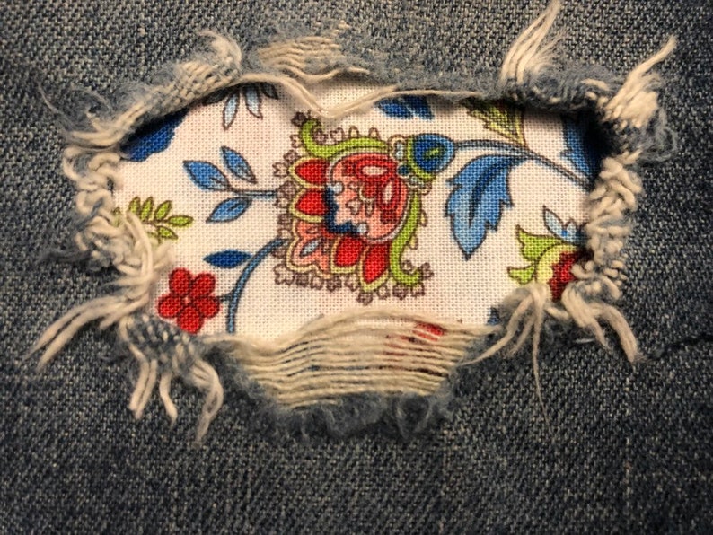 May include: A close-up of a pair of blue denim jeans with a hole that has been patched with a floral fabric. The fabric is white with a red, blue, and green floral pattern.