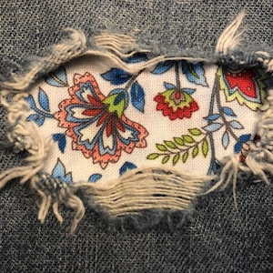 May include: A close-up of a ripped pair of blue jeans with a floral fabric patch sewn over the hole. The fabric patch features a white background with colorful floral designs in shades of blue, red, green, and pink.