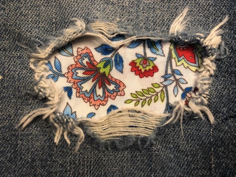 May include: A close-up of a ripped pair of blue jeans with a floral fabric patch sewn over the hole. The fabric patch features a white background with colorful floral designs in shades of blue, red, green, and pink.