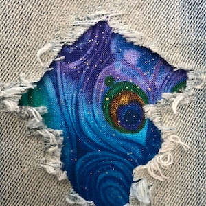 May include: A close-up of a ripped pair of blue jeans with a colorful peacock feather fabric patch sewn over the hole. The fabric patch has a blue, green, and purple swirl pattern with gold glitter accents.