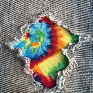 May include: A close-up of a ripped pair of blue jeans with a colorful tie-dye patch sewn over the hole.