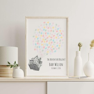 Puede incluir: Una impresi&oacute;n enmarcada con una ilustraci&oacute;n de una casa y globos de huellas dactilares de colores. El texto dice "The Adventure Begins!" y "Baby Wilson, October 17, 2026." La impresi&oacute;n est&aacute; en un estante blanco con una l&aacute;mpara y jarrones.