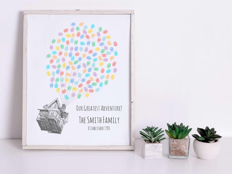 Puede incluir: Impresi&oacute;n enmarcada con una ilustraci&oacute;n de una casa y un globo de huellas dactilares de colores. El texto dice "Our Greatest Adventure! The Smith Family Established 1980." Tres peque&ntilde;as suculentas en macetas est&aacute;n frente a la impresi&oacute;n.