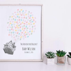 Puede incluir: Una impresi&oacute;n enmarcada con una ilustraci&oacute;n de una casa y un grupo de huellas dactilares de colores que se asemejan a un globo. El texto dice "The Adventure Begins!" e incluye un nombre y una fecha. Tres peque&ntilde;as suculentas en macetas est&aacute;n al frente.