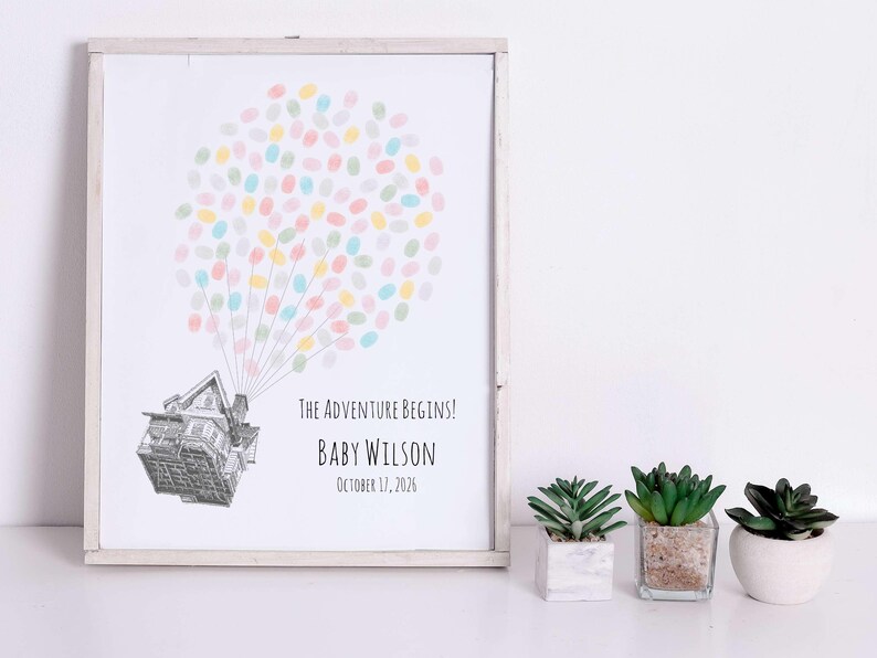 Puede incluir: Una impresi&oacute;n enmarcada con una ilustraci&oacute;n de una casa y un grupo de huellas dactilares de colores que se asemejan a un globo. El texto dice "The Adventure Begins!" e incluye un nombre y una fecha. Tres peque&ntilde;as suculentas en macetas est&aacute;n al frente.